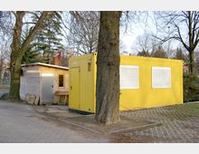 Gelbes Containergebäude mit geschlossenen Rollläden neben einem kleinen Schuppen aus Holz mit Flachdach, umgeben von Bäumen auf einem gepflasterten Platz. Im Hintergrund sind einige Büsche und Gebäude sichtbar.