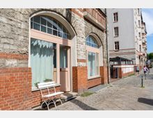 Fassade eines städtischen Wohngebäudes mit gemischtem Mauerwerk aus roten Ziegelsteinen und hellgrauem Naturstein. Im Erdgeschoss befinden sich große Fenster mit Rundbögen und rosa Holzrahmen, daneben steht eine weiße Bank. Der Gehweg davor ist mit Kopfsteinpflaster gestaltet, und im Hintergrund sind weitere Wohngebäude sowie Passanten zu sehen.