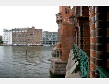 Backsteinfassade einer Brücke im Vordergrund mit grünen Metallgeländern, daneben ein Wasserkanal. Im Hintergrund stehen Gebäude unterschiedlicher Baustile, darunter ein modernes Glasgebäude und ältere Backsteingebäude.