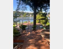 Holzterrasse mit Geländer und Topfpflanzen, im Vordergrund ein runder Holztisch. Dahinter Blick auf einen See mit Uferhäusern, umgeben von Bäumen.