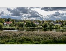 Dorfähnliche Landschaft mit einem ruhigen Teich im Vordergrund, umgeben von Schilfrohr und Bäumen. Im Hintergrund stehen Häuser mit roten und grauen Dächern in einer grünen Umgebung unter einem bewölkten Himmel.