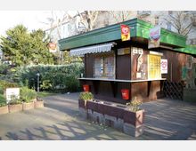 Ein kleiner Kiosk aus dunklem Holz mit grünem Flachdach und einer gestreiften Markise, umgeben von Pflanzen und einem gepflasterten Hof. Im Vordergrund stehen Blumenkübel aus Beton; im Hintergrund sind Bäume und mehrstöckige Wohnhäuser zu sehen.
