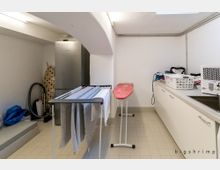 Hauswirtschaftsraum mit gefliesten Böden, einem Bügelbrett, einem Wäscheständer mit aufgehängter Kleidung, einem Kühlschrank, sowie einer Arbeitsfläche mit Waschbecken und Wäschekörben. Links stehen ein Staubsauger und weitere Haushaltsgegenstände.