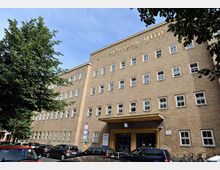 Backsteinfassade eines rechteckigen Gebäudes mit der Aufschrift „Stadtbad Mitte“ oberhalb der Fensterreihe. Im Vordergrund sind geparkte Autos, Fahrräder und ein Baum zu sehen.