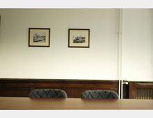 Ein schlichter Besprechungsraum mit weiß gestrichenen Wänden, zwei gerahmten Schwarz-Weiß-Zeichnungen an der Wand und einer dunklen Holzvertäfelung an der unteren Wandhälfte. Im Vordergrund steht ein Holztisch mit zwei gepolsterten Stühlen.