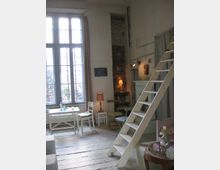 Ein heller Raum mit hohen Decken, großen Fenstern mit Sprossen und weißen Vorhängen. Der Boden besteht aus hellen Holzdielen, es gibt eine Holzleiter, einen kleinen Esstisch mit Stühlen, sowie eine gemauerte Wandnische mit Regal und Stehlampe.