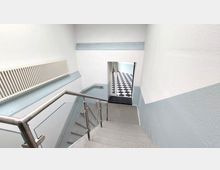 Treppenhaus mit hellem Wandanstrich, einem Metallhandlauf mit Glasfüllung und einem Heizkörper an der Wand. Am unteren Ende befindet sich ein Durchgang zu einem Raum mit schwarz-weiß kariertem Boden.