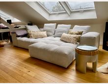 Helles Wohnzimmer unter einer Dachschräge mit mehreren Dachfenstern. Zentral steht ein großes, graues Sofa mit dekorativen Kissen, daneben ein kleiner Beistelltisch aus Metall und Holz, auf einem Holzboden.