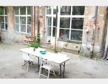 Ein gepflasterter Innenhof mit einem einfachen weißen Tisch und Stühlen im Vordergrund. Die umliegenden Gebäude haben sichtbare Backsteinfassaden, teilweise verputzt, mit großen Fenstern und Zeichen von Verwitterung.