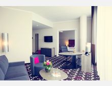 Wohnbereich eines modernen Hotelzimmers mit einem grauen Sofa, einem Sessel mit pinkem Kissen, einem runden Tisch mit Blumen und Gläsern sowie einem Schreibtisch mit Stuhl. Im Hintergrund ein Flachbildfernseher, ein dekorativer Teppich mit geometrischem Muster und ein angrenzender Schlafbereich mit Bett und Nachttischlampe.