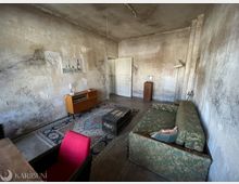 Verwittertes Wohnzimmer mit fleckigen, abblätternden Wänden und hohen Decken, eingerichtet mit einem grünen Sofa, einem gemusterten Teppich und einer dunklen Holztruhe als Couchtisch. An der Wand stehen ein niedriges Sideboard und ein kleiner Sockel, im Hintergrund führt eine weiße Tür in den nächsten Raum.