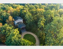 Luftaufnahme eines kleinen, klassizistischen Gebäudes mit Säuleneingang, umgeben von dichtem Wald. Vor dem Gebäude befindet sich ein runder, von einem Weg eingefasster Rasenplatz.