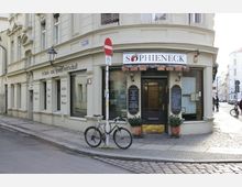 Eckhaus in heller Putzfassade mit einem Restaurant namens „Sophieneck“, dessen Eingang mit schwarzer Tür und Speisekarten an den Seiten versehen ist. Vor dem Gebäude stehen ein Fahrrad an einem Verkehrsschild und zwei Blumenkübel; links und rechts erstrecken sich gepflasterte Straßen.