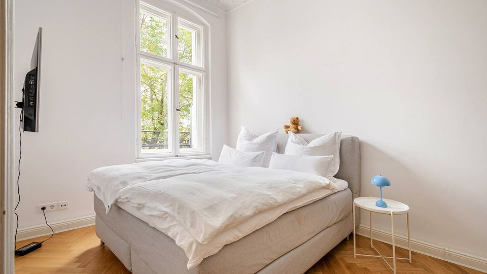 Helles Schlafzimmer mit großem Polsterbett und weißer Bettwäsche, neben einem hohen, zweiflügeligen Fenster mit Blick auf Bäume. Weiße Wände mit Stuckdecke, Fischgrätparkett sowie ein kleiner runder Beistelltisch mit blauer Lampe und ein an der Wand montierter Fernseher.