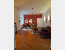 Wohnzimmer mit Parkettboden in Fischgrätmuster, einem roten Sofa rechts und einem niedrigen Couchtisch. Im Hintergrund ein Essbereich mit Tisch und Stühlen vor einer roten Wand, die mit einem Spiegel und Bildern dekoriert ist.