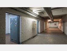 Ein einfacher Kellerflur mit weißen Ziegelwänden, grauem Betonboden und sichtbaren Lüftungsrohren an der Decke. Links ist eine offenstehende graue Tür mit Aufklebern, und an den Wänden sind Verteilerkästen angebracht.