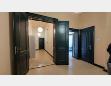 Ein Innenbereich mit beigefarbenen Wänden und dunklen Holztüren; ein Flur mit mehreren Türen auf beiden Seiten, leuchtende Kugellampen hängen an der Decke. Im Hintergrund ist ein weiterer Raum erkennbar.