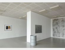 Minimalistischer Galerieraum mit grauem Boden und weißen Wänden. An den Wänden hängen ein Foto einer Person mit gelben Haaren, eine graue, abstrakte Zeichnung und eine weitere großflächige Arbeit; im Vordergrund steht ein silbernes Metallfass.