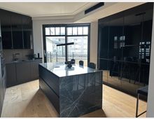 Moderne Wohnküche mit großer Kochinsel aus dunkel marmorierter Steinplatte und umlaufenden mattschwarzen Schrankfronten. Eine breite Fensterfront mit Balkontür bringt Tageslicht hinein; rechts sind hochglänzende, spiegelnde Schranktüren und Holzfußboden zu sehen.