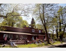 Backsteinbau mit Terrasse, umgeben von Bäumen und Wiesen, dient als Café oder Restaurant. Vor dem Gebäude stehen Sitzbänke, Sonnenschirme, und Liegestühle, die eine einladende Außenatmosphäre schaffen.