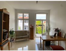 Helle Wohnzimmereinrichtung mit weißen Wänden, großen Fenstern und einem Balkon. Links steht ein Bücherregal, daneben ein Sofa, rechts ein Holztisch mit einem Laptop; verschiedene Pflanzen und Dekorationen verteilen sich im Raum.