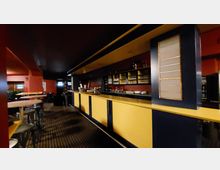 Barraum mit langen, schwarz-gelben Tresen im Vordergrund, dahinter Regalflächen mit Gläsern und Flaschen. Die Wände sind rot gestrichen, und die Decke sowie einige Säulen in dunklen Farben gehalten. Links stehen runde Tische mit Hockern auf einem gemusterten Teppichboden.