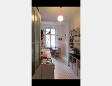 Schmale Küche mit weiß gefliesten Boden und schwarzen Punkten, links eine Arbeitsfläche mit Herd und Spüle, rechts offene Regale mit Küchenutensilien. Im Hintergrund ein kleiner Essbereich mit Holztisch, roten Stühlen und einem großen Fenster, das Tageslicht hereinlässt.