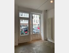 Ein leerer Raum mit großen Glasfenstern und einer Glastür, die auf eine belebte Straße mit parkenden Autos und Fahrrädern blickt. An der rechten Wand hängt ein vertikaler Heizkörper, der Boden ist aus Beton.