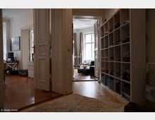 Flur in einer Wohnung mit Holzboden, Teppich und einem großen Regal mit Schuhen an der rechten Wand. Durch eine geöffnete Tür sind ein Raum mit Polstermöbeln, einem großen Fenster und Vorhängen zu sehen.