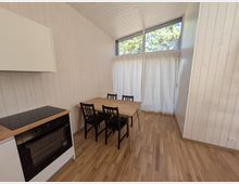 Heller Essbereich in einem schmalen Raum mit weißen Holzvertäfelungen, Holzfußboden und einem kleinen Tisch mit vier Stühlen neben einer Küchenzeile. Am Ende des Raums befindet sich eine bodennahe Fensterfront mit Vorhängen und einem hohen Oberlichtfenster mit Blick auf Bäume.