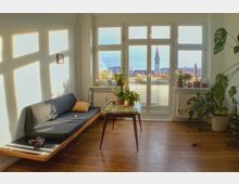 Wohnzimmer mit hellem Holzboden, einem grauen Sofa links, einem Glastisch mit Pflanzen in der Mitte und großen Fenstern, die auf einen Balkon und eine Stadtlandschaft mit einem Kirchturm im Hintergrund blicken. Zimmerpflanzen stehen am rechten Rand und auf der Fensterbank.