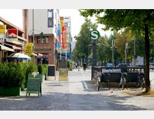Straßenszene mit Gehweg, gesäumt von Cafés, Restaurants und Schildern, darunter eine Beschilderung der S-Bahn ("S" für Anhalter Bahnhof); daneben eine Fahrradabstellanlage und eine baumbestandene Allee im Hintergrund.