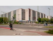 Mehrstöckiges Wohngebäude aus Beton mit Balkonen, teils in grünen und roten Farbakzenten, umgeben von Gehwegen, Bäumen und einer Straßenkreuzung mit Ampeln. Im Erdgeschoss befinden sich Geschäfte, darunter ein Fitnessstudio.