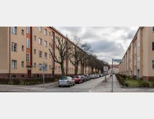 Straßenszene mit Wohngebäuden aus der Nachkriegszeit, gekennzeichnet durch helle Putzfassaden und rotes Klinkermauerwerk im Sockelbereich. Die Straße ist gepflastert, gesäumt von kahlen Bäumen, parkenden Autos und einem Gehweg mit kleinen Grünflächen; Straßenschilder weisen auf die Granitzstraße hin.