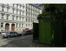 Straßenszene mit Wohnhäusern im historischen Baustil, großen Fenstern und verzierten Fassaden. Im Vordergrund steht ein grüner, gusseiserner Pavillon, möglicherweise eine historische öffentliche Toilette, umgeben von parkenden Autos und Bäumen.