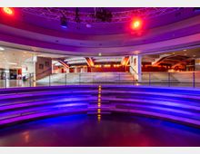Halbrunder Veranstaltungsraum mit terrassenförmigen Sitzreihen und moderner Beleuchtung in violetten und roten Farben. Im Hintergrund befindet sich ein Treppenaufgang, der zu einer oberen Etage mit weitläufiger Architektur führt.