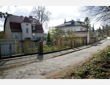 Straßenansicht mit zwei Wohnhäusern aus der ersten Hälfte des 20. Jahrhunderts, umgeben von kleinen Gärten, die durch Metallzäune mit Backsteinpfosten abgegrenzt sind. Die Straße ist asphaltiert und von vereinzelten Bäumen und Sträuchern gesäumt.