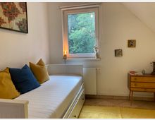 Schlicht eingerichtetes Zimmer mit einem weißen Tagesbett, dekoriert mit blauen und gelben Kissen. Im Hintergrund befindet sich ein Fenster mit Blick auf grüne Bäume, daneben eine kleine Holzkommode mit Büchern und Dekoration.