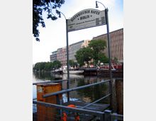 Historischer Hafen in Berlin mit mehreren festgemachten Booten am Wasser. Im Vordergrund ein Steg mit Schild, im Hintergrund moderne Gebäude und Bäume entlang des Ufers.