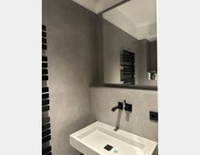 Modernes Badezimmer mit grauen Wandfliesen, rechteckigem weißem Waschbecken und mattschwarzer Wandarmatur. Darüber hängt ein großer Spiegel mit dunklem Rahmen; am Rand ist ein schwarzer Handtuchheizkörper zu sehen.