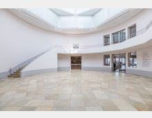 Heller, runder Museumsraum mit einer gewölbten Wand, großen Fenstern aus Glasbausteinen im oberen Bereich und einem offenen Zugang zu weiteren Räumen. Der Boden besteht aus hellen, geometrisch angeordneten Fliesen, und an der linken Seite führt eine geschwungene Treppe nach oben.