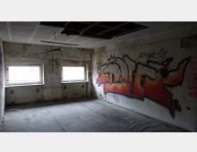 Ein verlassenes und heruntergekommenes Gebäude mit beschädigten Wänden, zwei kleinen Fenstern und einer großen Graffiti-Malerei in orange, rot und gelb an einer der Wände. Die Decke weist Löcher auf, und der Raum ist ohne Möbel.