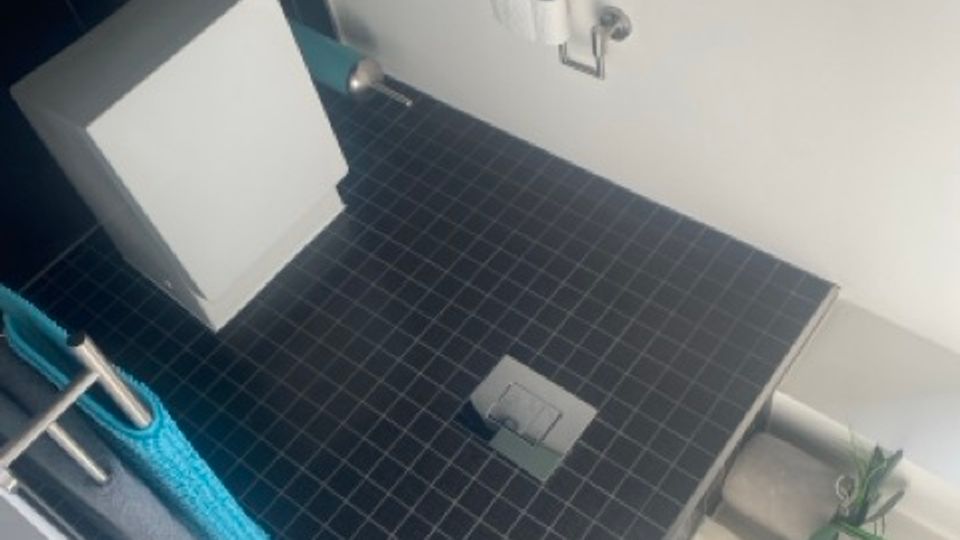Blick von oben in ein kleines, gefliestes Badezimmer mit dunklen Bodenfliesen, bodengleicher Dusche mit Ablaufrinne und Glasabtrennung. An der Wand sind eine Toilettenpapierhalterung und eine kompakte weiße Sanitärinstallation zu sehen, daneben ein Handtuchhalter mit blauem Handtuch.