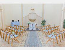 Heller Kirchenraum mit einem schlichten Altarbereich, dekoriert mit Blumenarrangements. Holzstühle mit hellblauen Polstern sind in Reihen aufgestellt, ein Mittelgang mit Teppich führt zum Altar, flankiert von Kerzen und Grünpflanzen.