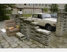 Wohnhof mit Betonplattenbelag, einer unvollständigen gemauerten Steinmauer, einem abgenutzten Sessel und einem geparkten, beigen Auto; im Hintergrund ein mehrstöckiges Wohngebäude mit verputzter Fassade und Fenstern.