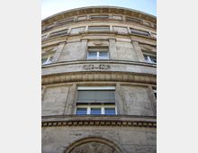 Fassade eines historischen Gebäudes aus Naturstein mit vertikalen und horizontalen Zierelementen. Sicht auf mehrere Etagen mit Fenstern, teilweise mit geschlossenen Rollläden, sowie dekorativen Ornamenten oberhalb und unterhalb der Fenster.