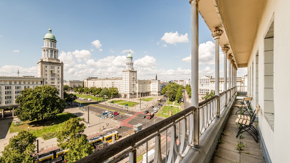 Blick von einem langen Balkon mit Säulen und Metallgeländer über eine breite Stadtstraße mit Straßenbahn, Kreuzung und Grünflächen. Gegenüber stehen zwei markante, mehrstöckige Gebäude mit runden Türmen und grünen Kuppeln vor blauem Himmel.