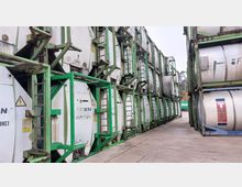 Gestapelte Industriegasbehälter in Metallrahmen, die in mehreren Reihen auf einem Lagerplatz aufgestellt sind, mit grün gestrichenen Stahlrahmen und einem Betonboden. Im Hintergrund sind Bäume und ein weiterer Behälter sichtbar.