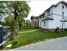 Einfamilienhaus im Landhausstil mit weißer Fassade und roten Klinkerelementen, umgeben von einem gepflegten Garten mit Rasen, Bäumen und Gehweg. Im Vordergrund ein schwarzes Metalltor und eine gepflasterte Auffahrt.