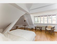 Dachzimmer mit weiß gestrichenen Wänden und Holzboden, einem Bett im Vordergrund, mehreren Sitzmöbeln aus Holz und Rattan sowie einem großen Fenster mit Blick auf Bäume. Die schräge Decke und sichtbare Dachbalken betonen die Dachgeschossarchitektur.